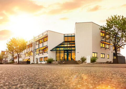 Zu sehen ist der Hauptsitz der wob AG in Viernheim. Die Kamera befindet sich nah am Boden und richtet ihren Fokus von unten auf ein großes, weißes, dreistöckiges Gebäude, dessen Fenster gelblich schimmern. Eine große Eingangstür aus Glas ist über ein paar Treppenstufen zu erreichen. Über dem Gebäude scheint hell die Sonne. | © wob AG