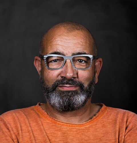 Dieses Bild ist ein Porträt von Roland Rudolf. Er hat einen Bart und eine Brille. Er trägt ein orangenes Sweatshirt. Der Hintergrund ist dunkel. | © wob AG