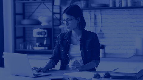 A young, casually dressed woman with short dark hair and glasses is sitting at a table and working on something on a laptop. A small office kitchen can be seen blurred in the background. There is a blue filter over the picture. | © wob AG