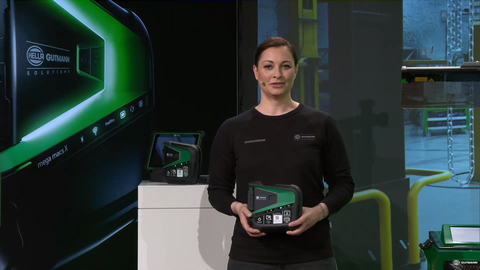 This image shows a screenshot from the digital live event. You can see presenter Lina van de Mars hosting the digital live event #Zuhausmesse. She is wearing a black long-sleeved shirt with her hair tied back at the nape of her neck and is looking into the camera. She is holding a mega macs X in her hands. Behind her is a presenter's desk and a large screen with the main campaign motif, a high-quality product image on a black background.  