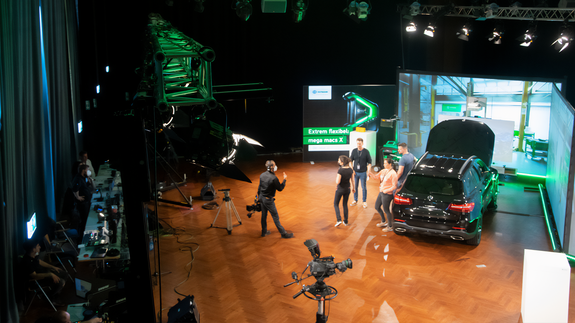 This picture shows the studio of the Zuhausemesse workshop show, including the camera setup. There is a car in the centre of the studio and two large screens behind it. Five people are standing in the studio.