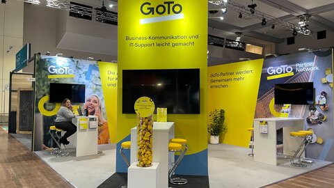 Das Foto zeigt einen Messestand von GoTo. Das ist daran zu erkennen, dass sich das Logo auf allen Messewänden widerspiegelt. Eine Dame sitzt an einem Tisch. Zu sehen sind noch weitere Tische, die Gesprächssituationen am Messestand erlauben sollen.