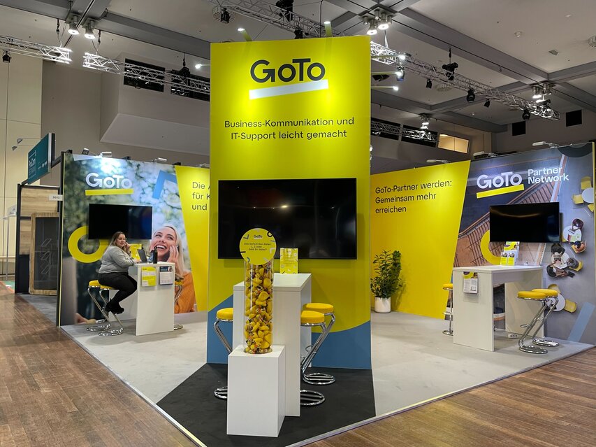 Das Foto zeigt einen Messestand von GoTo. Das ist daran zu erkennen, dass sich das Logo auf allen Messewänden widerspiegelt. Eine Dame sitzt an einem Tisch. Zu sehen sind noch weitere Tische, die Gesprächssituationen am Messestand erlauben sollen.