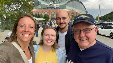 Auf einem Foto sind 4 Personen zu sehen, die fröhlich in die Kamera lächeln. Darunter sind zwei junge Frauen, ein junger Mann und ein etwas älterer Herr mit Baseballkappe. Hinter den vier Personen ist ein Baseballstadion zu erkennen.