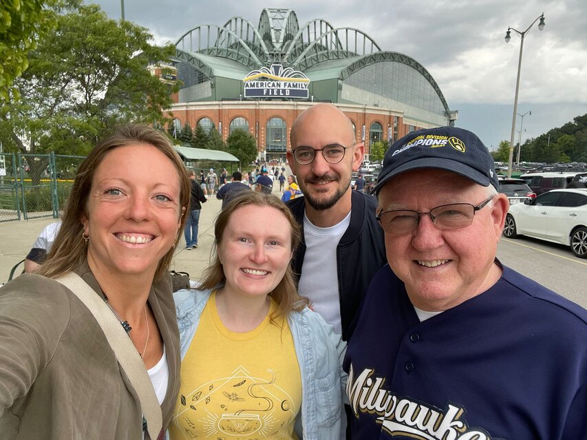 Auf einem Foto sind 4 Personen zu sehen, die fröhlich in die Kamera lächeln. Darunter sind zwei junge Frauen, ein junger Mann und ein etwas älterer Herr mit Baseballkappe. Hinter den vier Personen ist ein Baseballstadion zu erkennen.